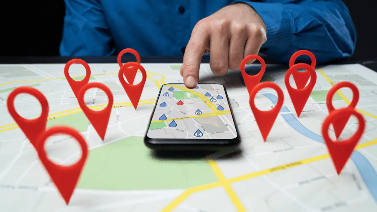 A hand pointing at a mobile phone showing a map with multiple red location pins on it.